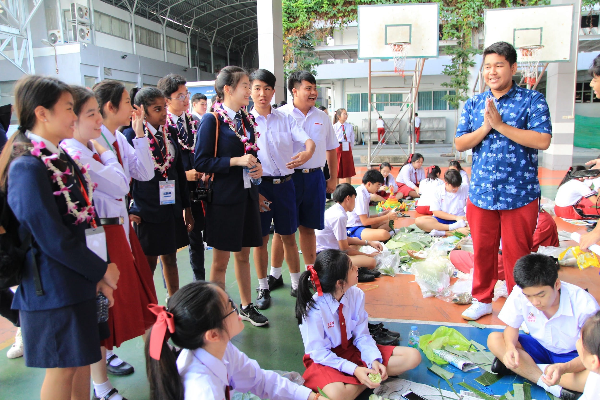 Students in school uniforms gather outdoors; some stand with flower leis while others sit on the ground working with leaves.